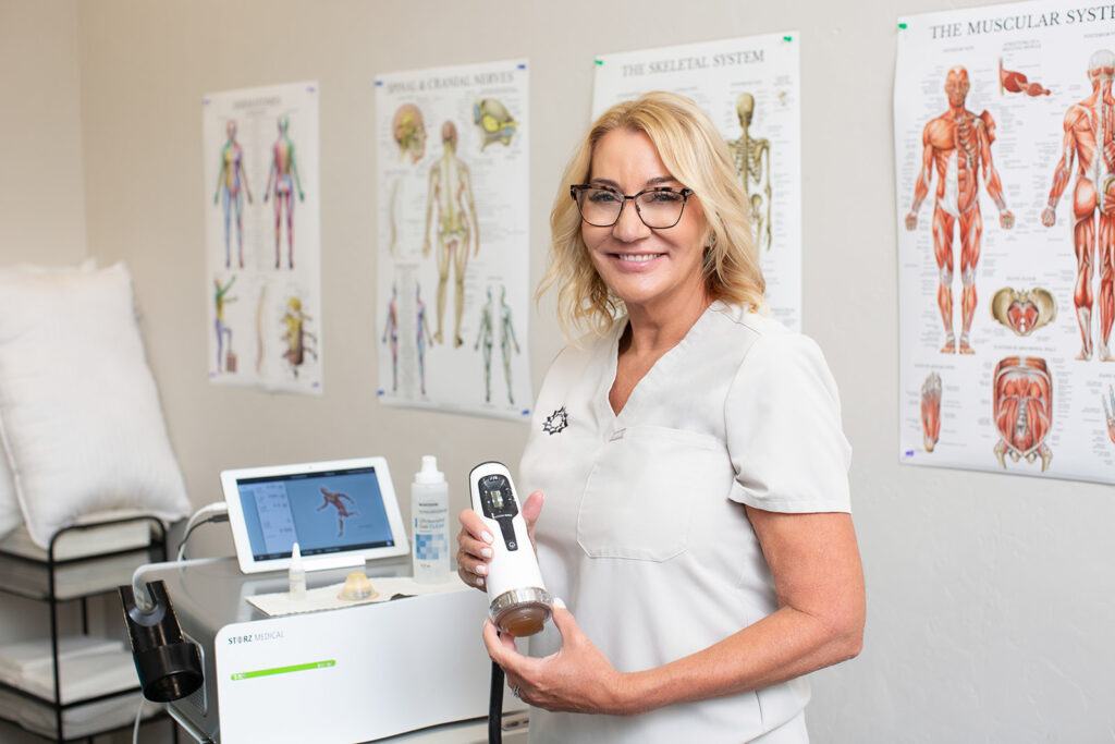 aesthetician holding a shock wave unit and looking at the camera with anatomy posters on the wall behind her